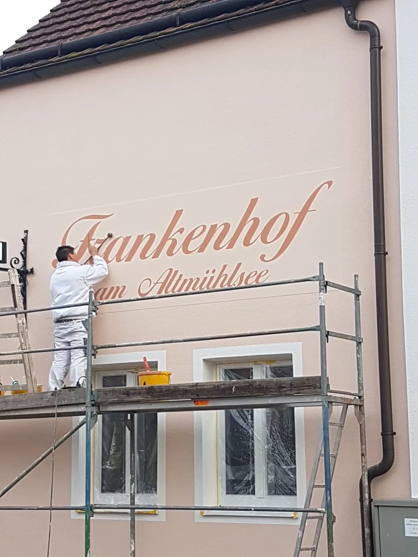 Maler bringt den Schriftzug „Frankenhof am Amühlsee“ an einer frisch gestrichenen Hausfassade mit Gerüst an.